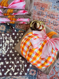 A styled flat-lay of folded fabric items on a patterned rug, including a brown tote bag with white polka dots and animal silhouettes, a brown fabric item with a pink and orange floral print, and a brown fabric item featuring a large orange-and-white gingham pumpkin design with a pink bow and gold rings on top.