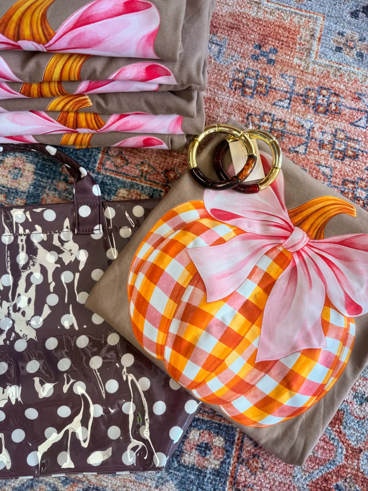 A styled flat-lay of folded fabric items on a patterned rug, including a brown tote bag with white polka dots and animal silhouettes, a brown fabric item with a pink and orange floral print, and a brown fabric item featuring a large orange-and-white gingham pumpkin design with a pink bow and gold rings on top.